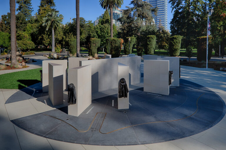 Circular stone memorial with engraved walls and bronze firefighter jackets in a landscaped city park.