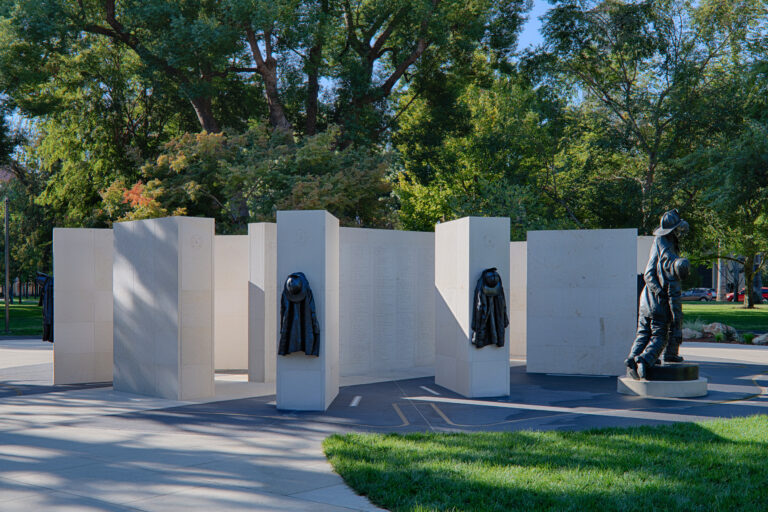 Firefighter memorial with white stone walls, empty jackets on pillars, and bronze firefighter statues in a park.