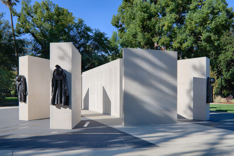 Outdoor memorial with tall stone walls engraved with names and hanging firefighter coats and helmets among trees