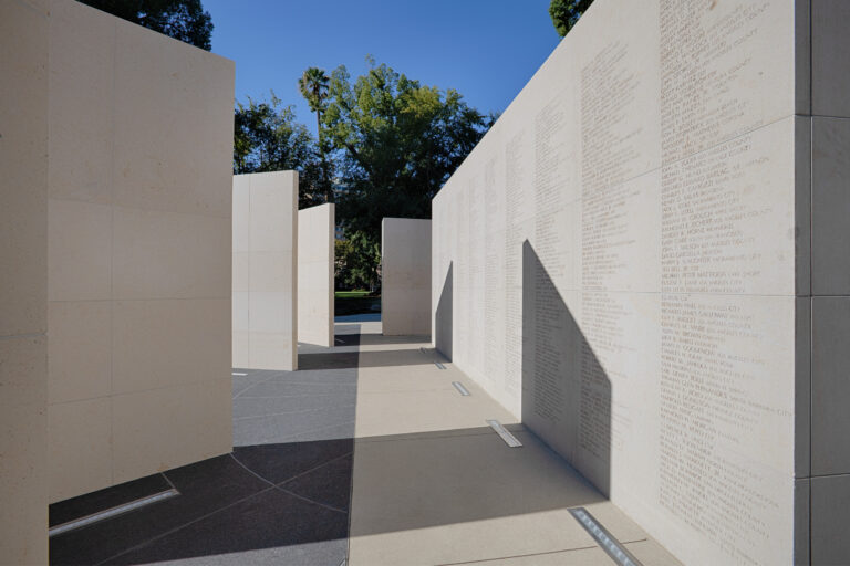 Walkway between tall pale stone memorial walls engraved with many names, with trees and blue sky beyond.