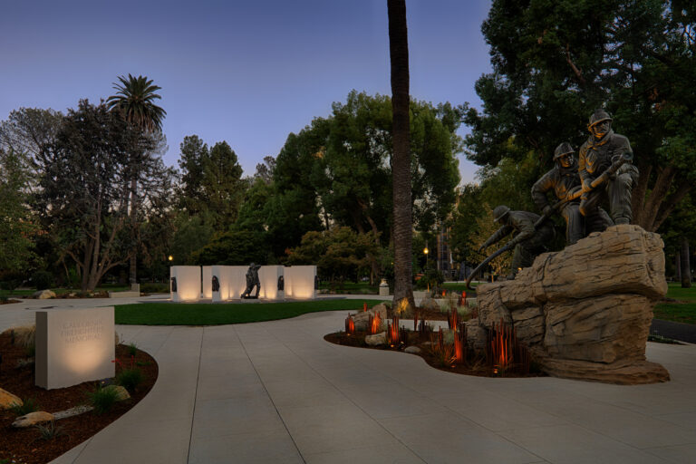 California Firefighters Memorial at dusk, with bronze firefighter statues, curved walkway, and illuminated walls in a park.