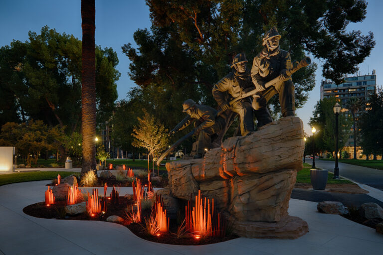 Illuminated statue of firefighters on a rock in a park at dusk, surrounded by glowing red memorial lights.