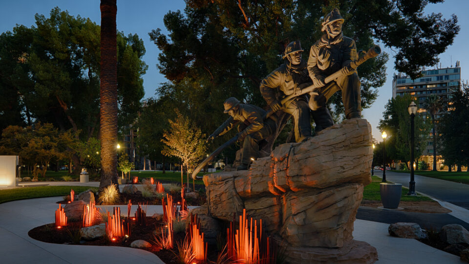 Illuminated statue of firefighters on a rock in a park at dusk, surrounded by glowing red memorial lights.