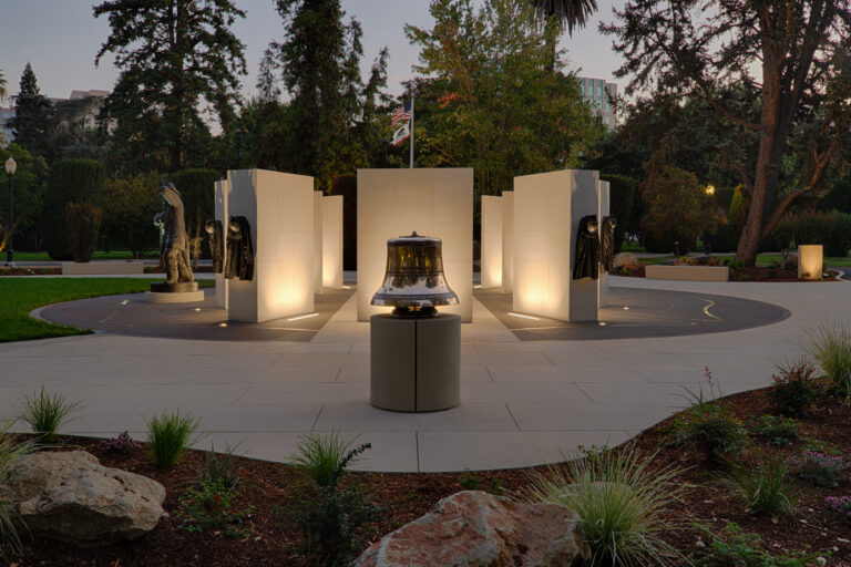 Illuminated memorial with a bell, firefighter statues and gear, and U.S. and California flags in a landscaped park at dusk