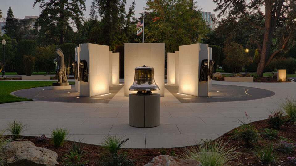 Illuminated memorial with a bell, firefighter statues and gear, and U.S. and California flags in a landscaped park at dusk
