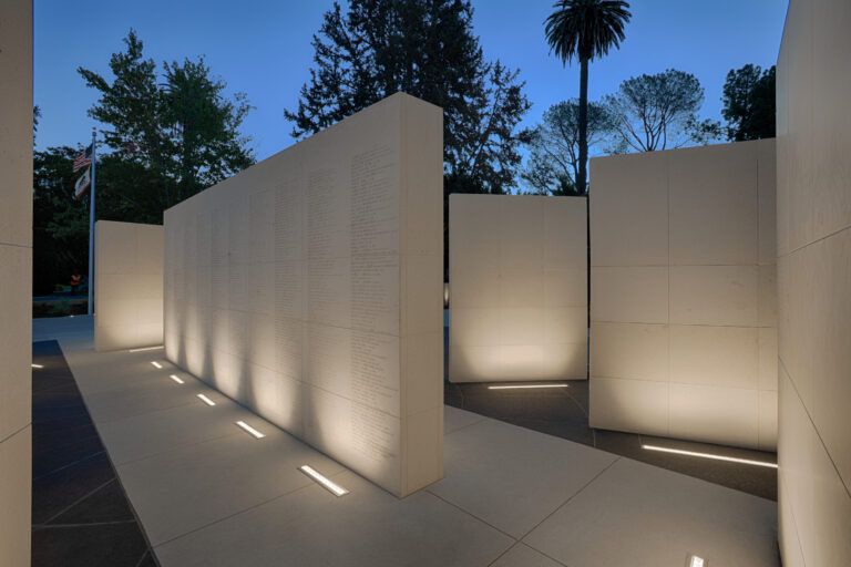 Illuminated stone memorial walls engraved with names at dusk, with trees and U.S. and California flags in the background