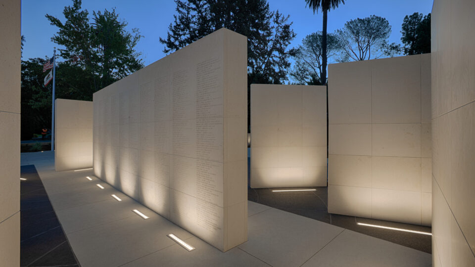 Illuminated stone memorial walls engraved with names at dusk, with trees and U.S. and California flags in the background