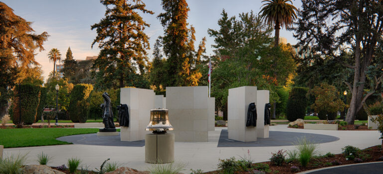 Memorial plaza in a landscaped park with stone pillars, bronze firefighter figures, and a bell at center.