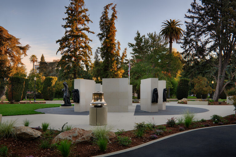 Memorial plaza in a landscaped park with stone pillars, bronze firefighter figures, and a bell at center.