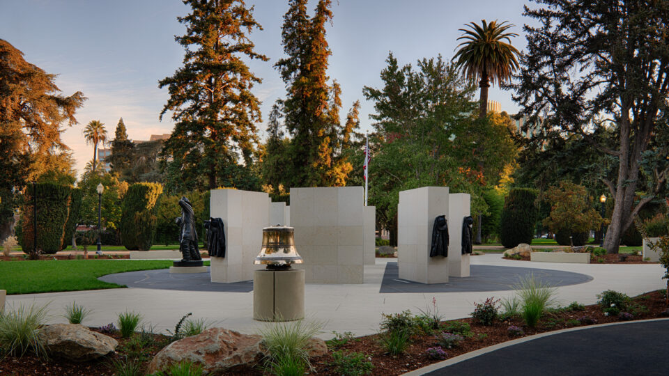 Memorial plaza in a landscaped park with stone pillars, bronze firefighter figures, and a bell at center.