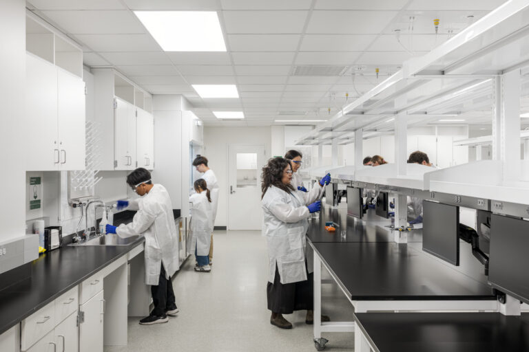 Researchers in white lab coats and blue gloves working at benches in a bright modern laboratory