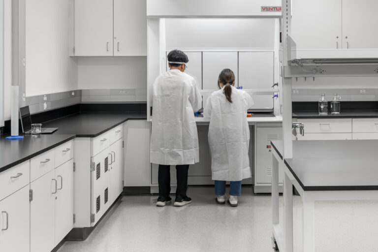 Two people in white lab coats working at a fume hood in a clean, modern laboratory.