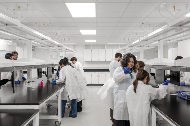 Students in white lab coats and gloves working at benches in a bright, modern laboratory.