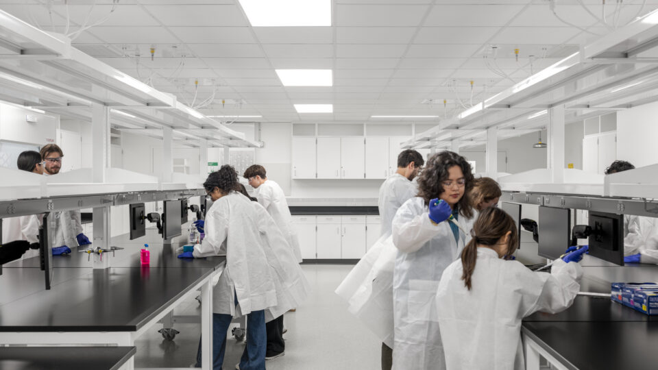 Students in white lab coats and gloves working at benches in a bright, modern laboratory.
