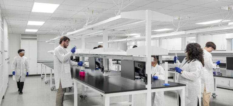 Researchers in white lab coats and blue gloves work at benches in a bright, modern laboratory.