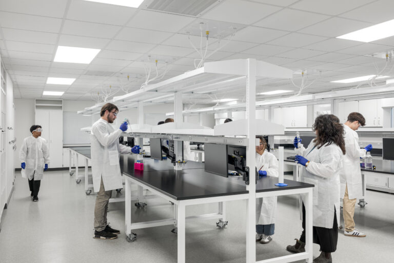 Researchers in white lab coats and blue gloves work at benches in a bright, modern laboratory.