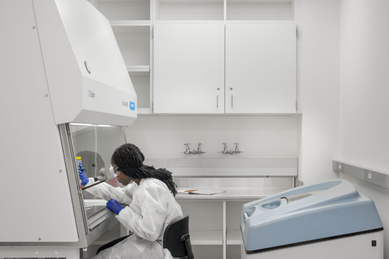 Scientist in protective gear working at a biosafety cabinet in a clean white laboratory.