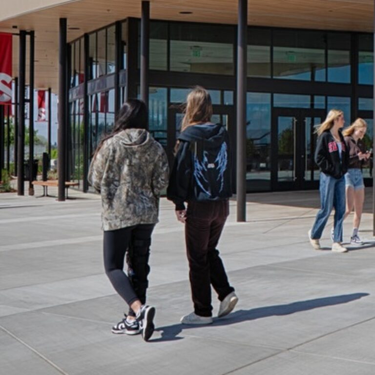 Two students walk toward a modern school entrance while two others stand talking nearby.