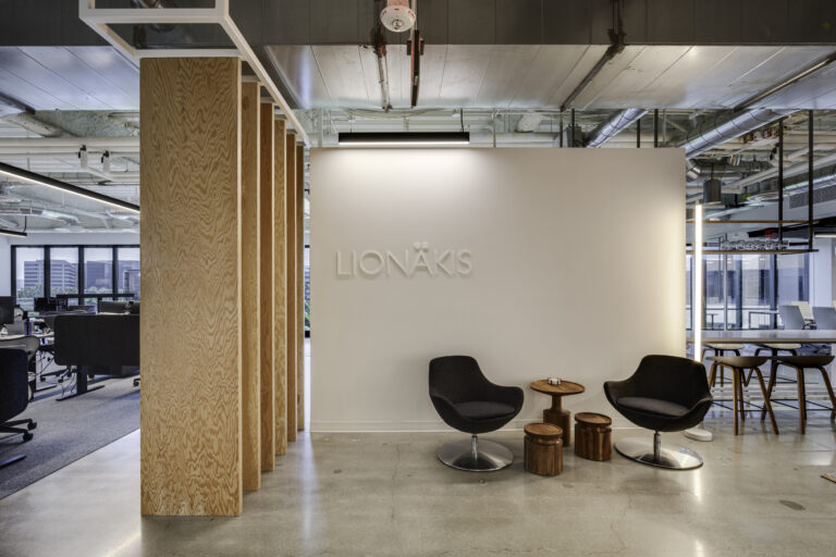Modern office lounge with two black chairs, small wooden tables, and a white wall displaying the Lionakis logo.