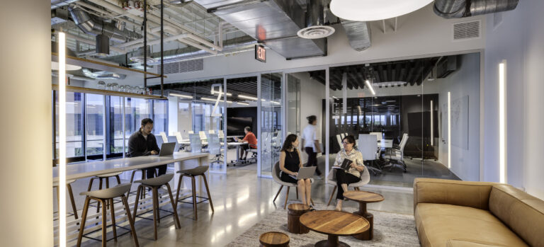 Modern office lounge with glass meeting rooms, exposed ductwork, tan sofa, and people working and talking.