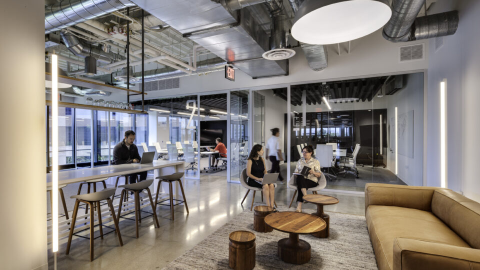 Modern office lounge with glass meeting rooms, exposed ductwork, tan sofa, and people working and talking.