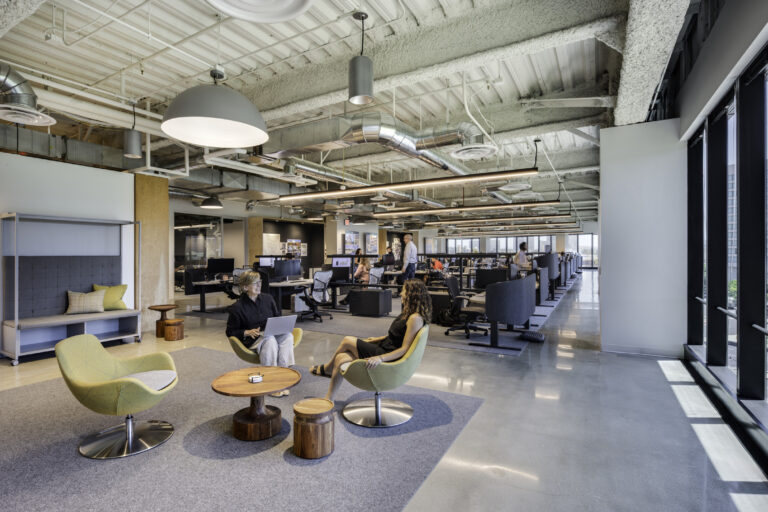 Modern open-plan office with exposed ceiling ducts, large windows, and two people talking in a lounge area.