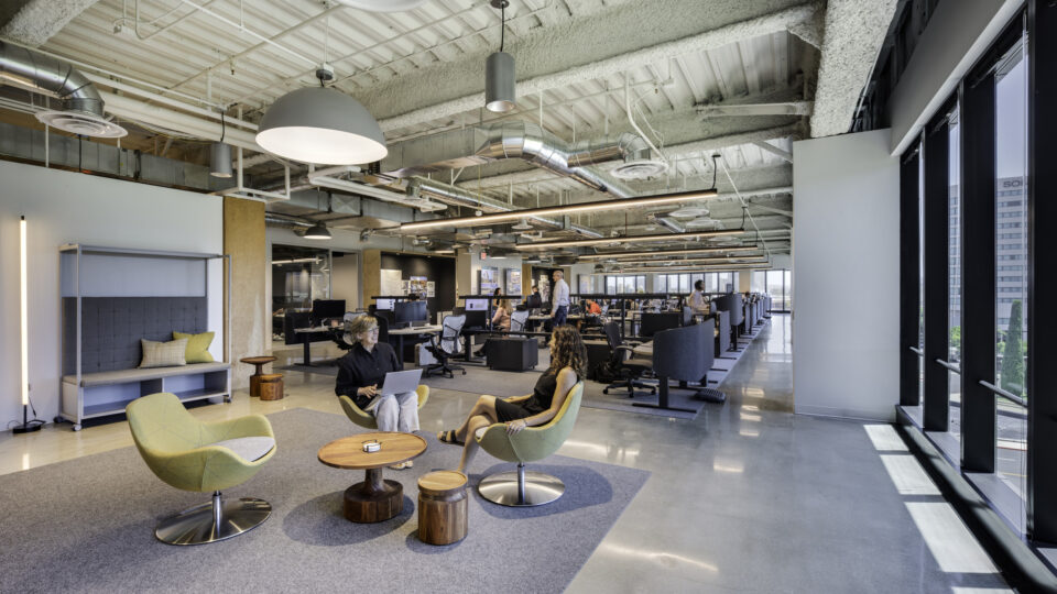 Modern open-plan office with exposed ceiling ducts, large windows, and two people talking in a lounge area.