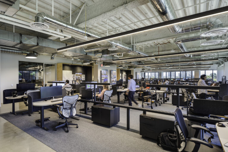 Modern open-plan office with rows of desks, computers, and workers beneath exposed ductwork and linear ceiling lights