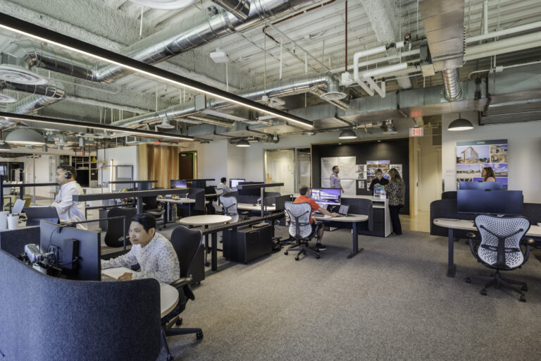 Open-plan office with employees at desks, exposed ductwork, and a small group talking near a presentation wall