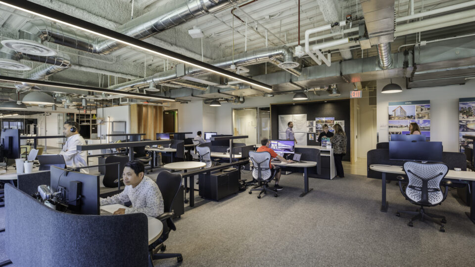Open-plan office with employees at desks, exposed ductwork, and a small group talking near a presentation wall