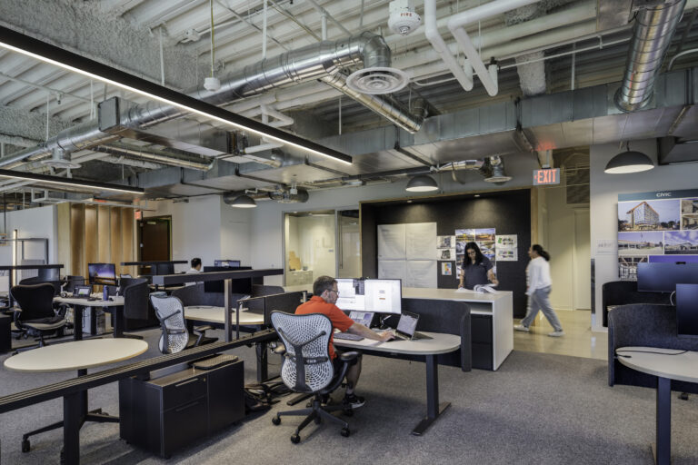 Open-plan office with desks, computer monitors, and people working beneath an exposed industrial ceiling.
