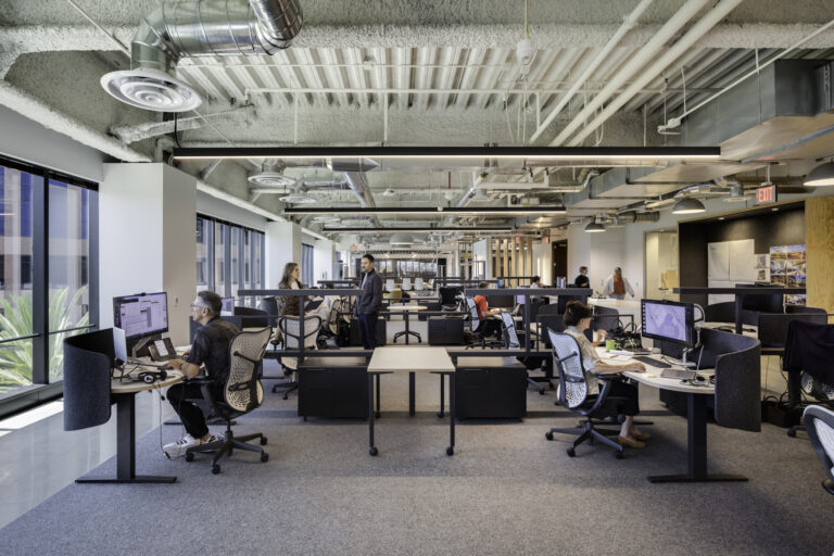 Open-plan office with several people working at computers beneath an exposed industrial ceiling and large windows.