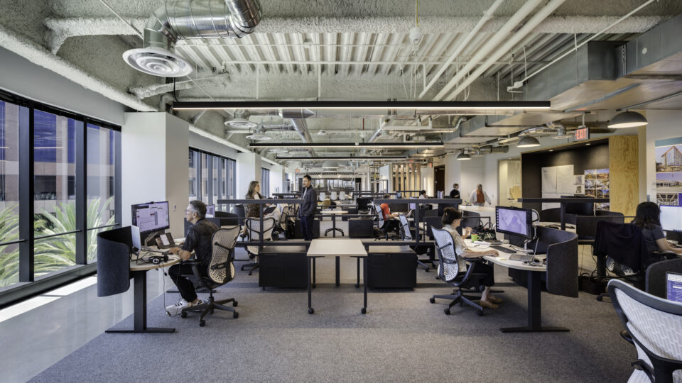 Open-plan office with several people working at computers beneath an exposed industrial ceiling and large windows.