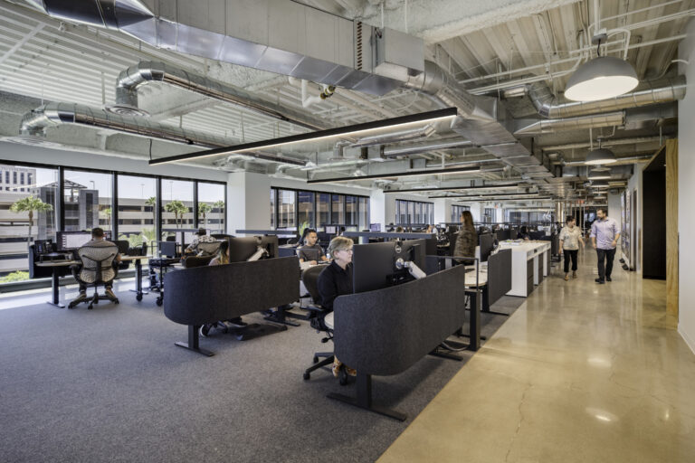 Open-plan office with employees at computer workstations, large windows, and exposed industrial ductwork overhead.