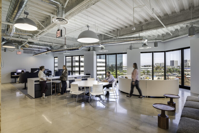 Open-plan modern office with exposed ductwork, round meeting table, lounge seating, and people working by large windows.