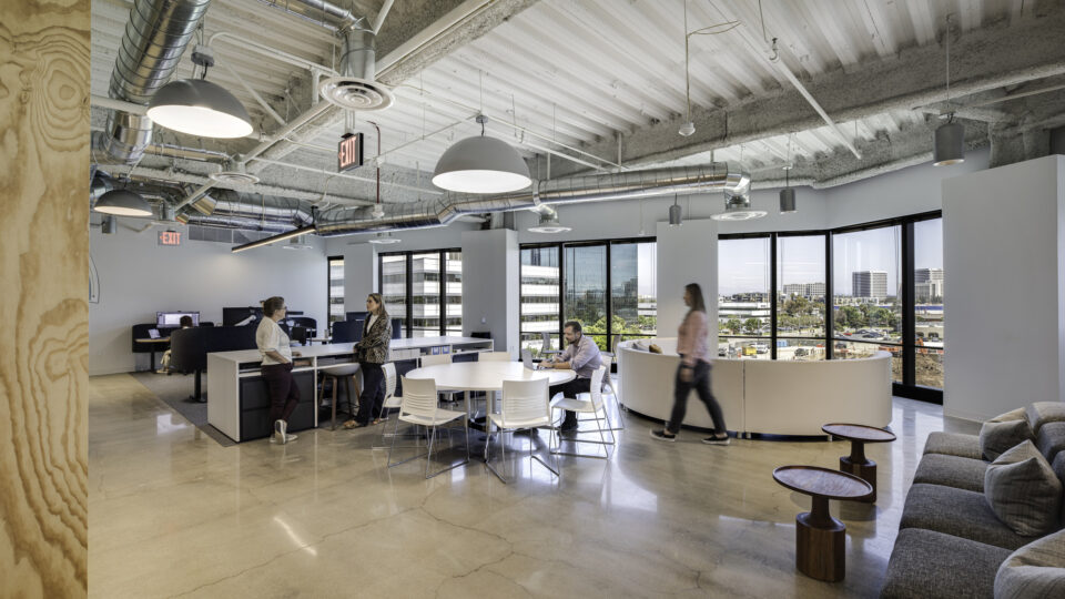 Open-plan modern office with exposed ductwork, round meeting table, lounge seating, and people working by large windows.