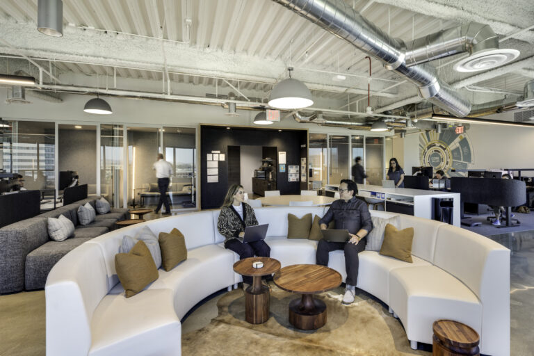 Modern open-plan office with coworkers at desks and two people with laptops seated on a curved white sofa.