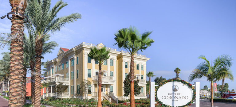 Hotel del Coronado sign beside a palm-lined yellow building and landscaped walkway under a clear blue sky