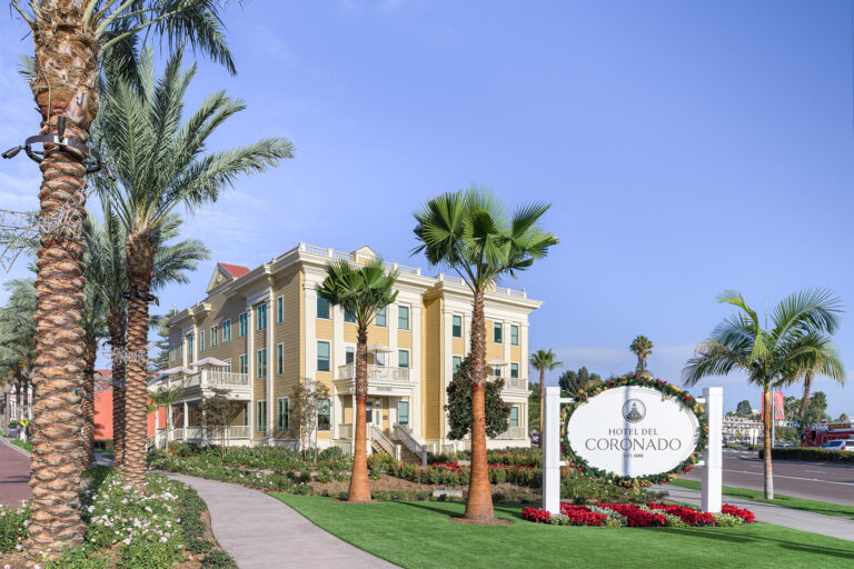 Hotel del Coronado sign beside a palm-lined yellow building and landscaped walkway under a clear blue sky
