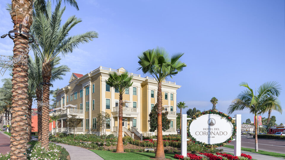 Hotel del Coronado sign beside a palm-lined yellow building and landscaped walkway under a clear blue sky