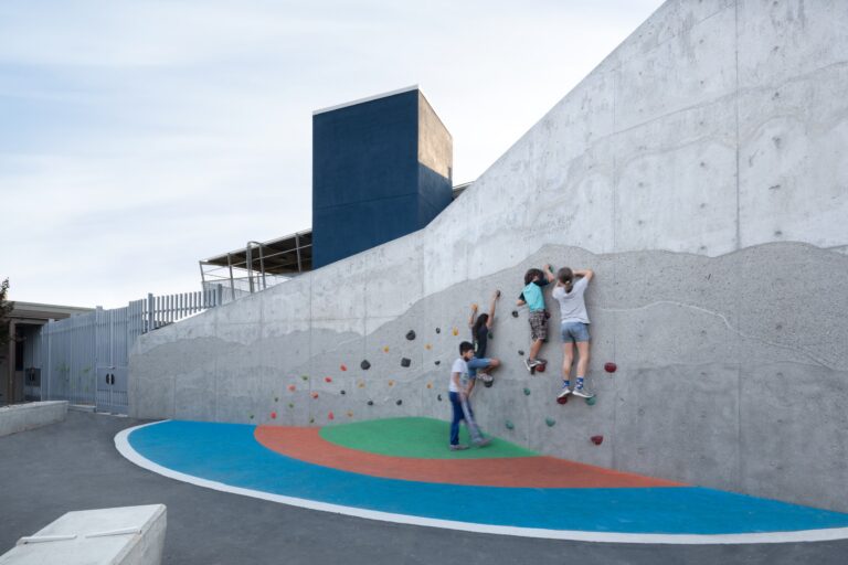 Children climbing a concrete play wall with colorful handholds beside a modern building and painted ground.