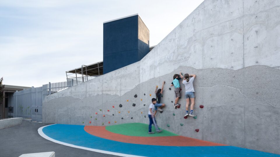 Children climbing a concrete play wall with colorful handholds beside a modern building and painted ground.