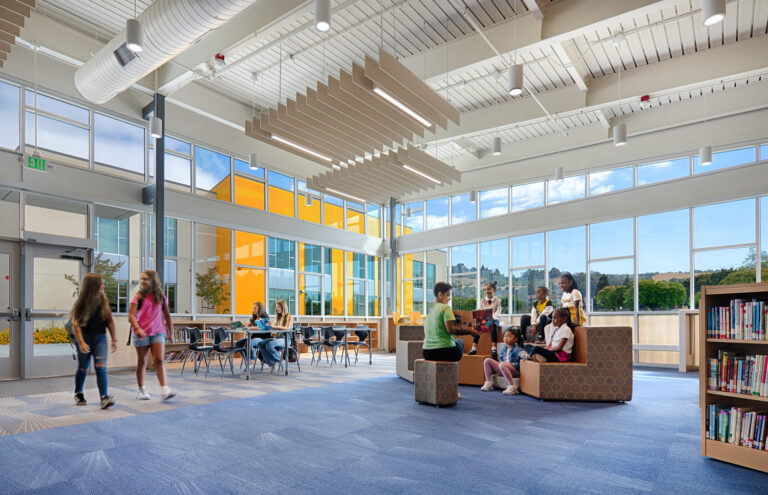 Bright modern school library with large windows, bookshelves, tables, and children reading and walking inside