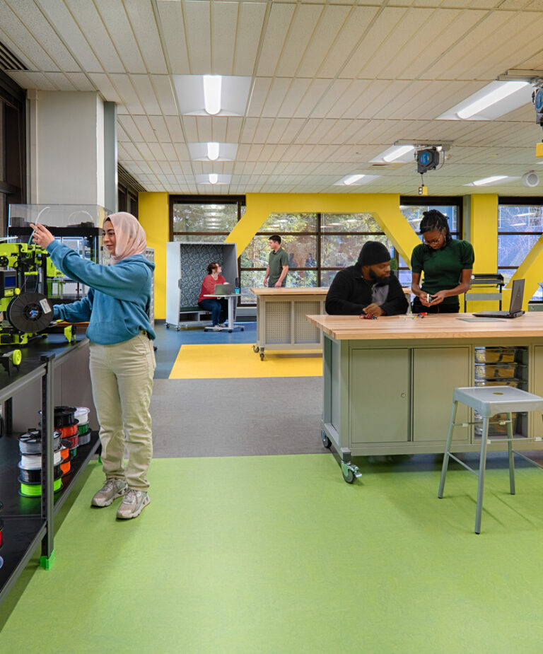 Students working in a bright makerspace with 3D printers, worktables, laptops, and a whiteboard.