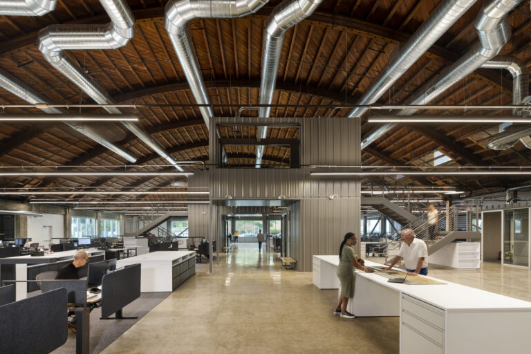 Open-plan industrial office with exposed wooden ceiling beams, large silver ducts, desks, and two people at a counter