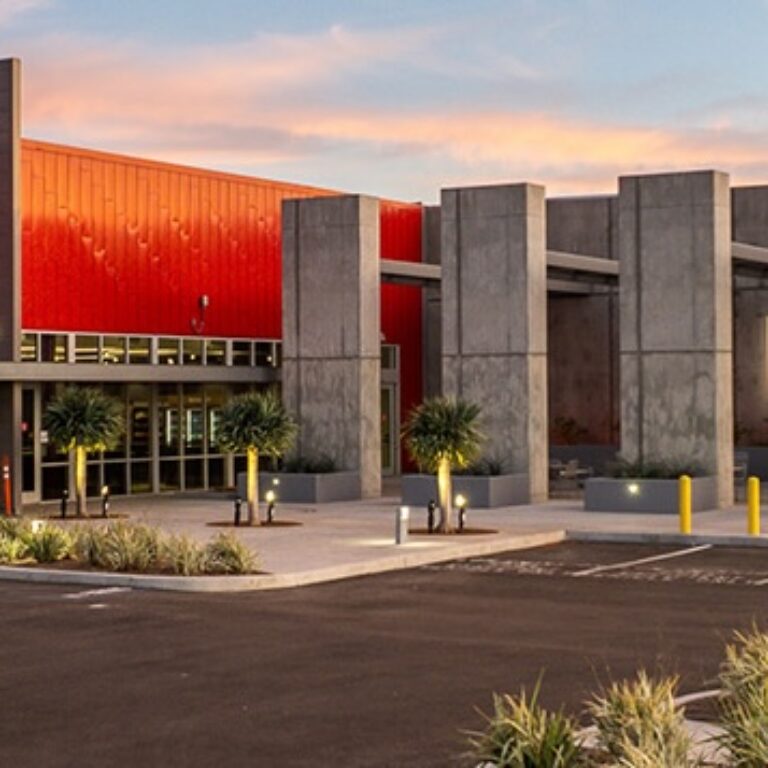 Modern building with a red metal facade and tall concrete columns at dusk, viewed from an empty parking lot