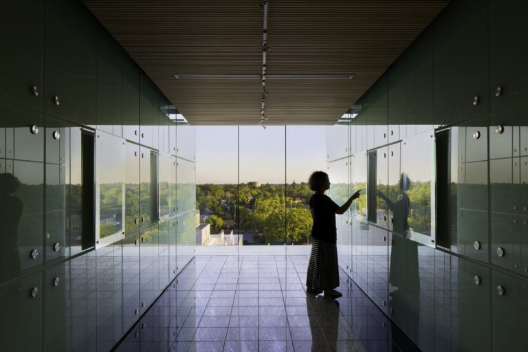 Silhouetted person touching a glass wall in a modern hallway with reflective panels and a tree-filled view outside