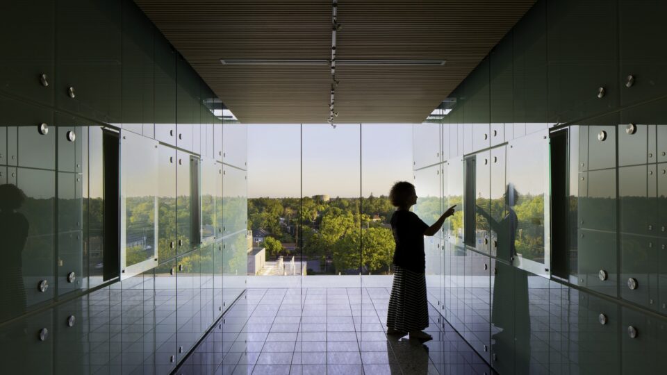 Silhouetted person touching a glass wall in a modern hallway with reflective panels and a tree-filled view outside
