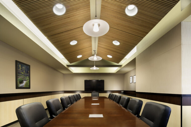 Modern conference room with a long wooden table, black chairs, a wall-mounted TV, and a slatted wood ceiling.