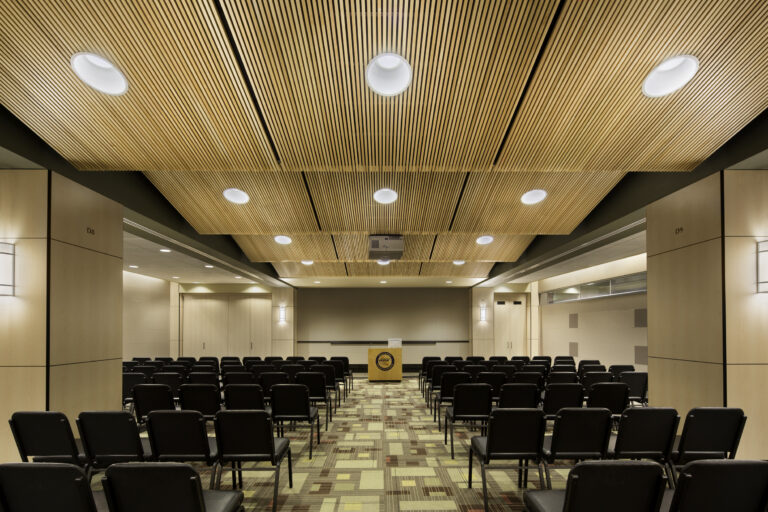 Empty conference room with rows of black chairs, a podium, and a wood slat ceiling with recessed lights.